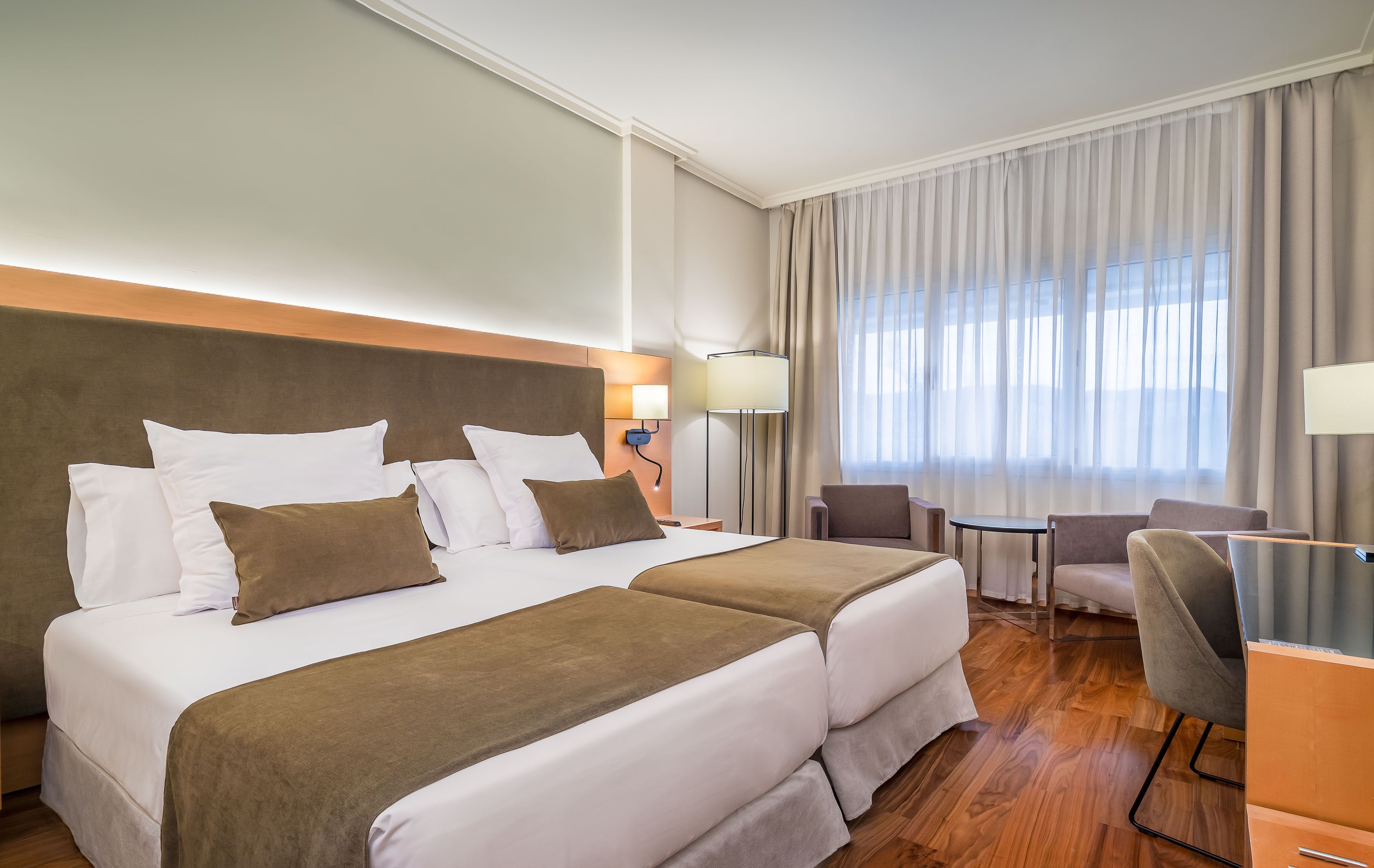 A hotel room with two beds, a desk, a chair, and a window with curtains.