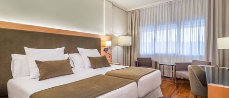 Habitación de hotel con dos camas, un escritorio, una silla y un ventanal con cortinas.