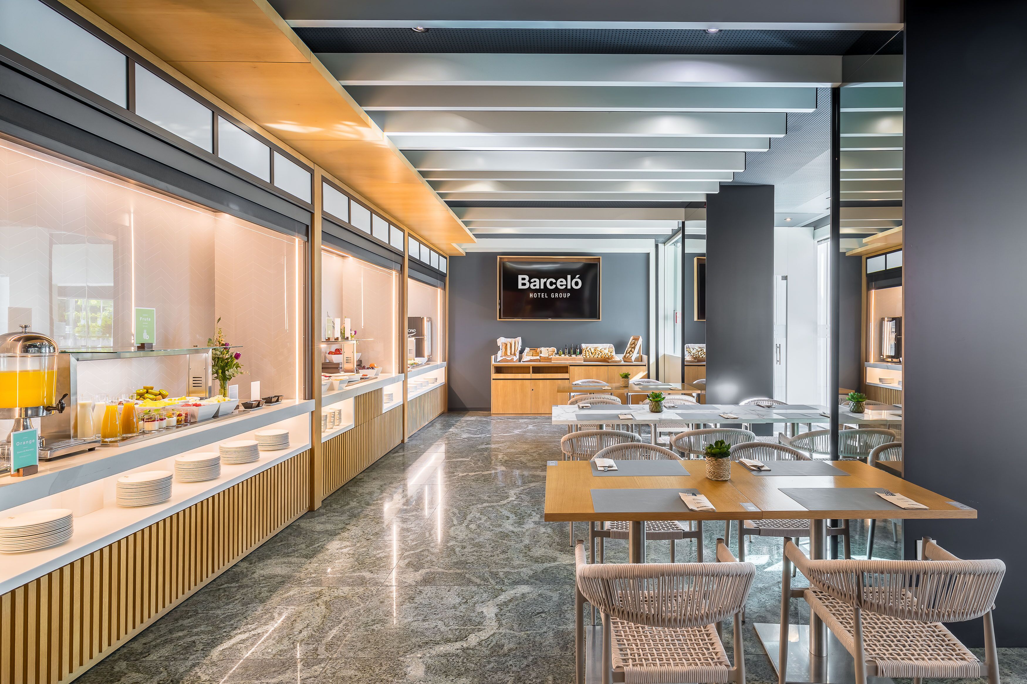 Un moderno vestíbulo de hotel con barra de desayuno, zona de estar y cartel del Barceló Hotel Group.