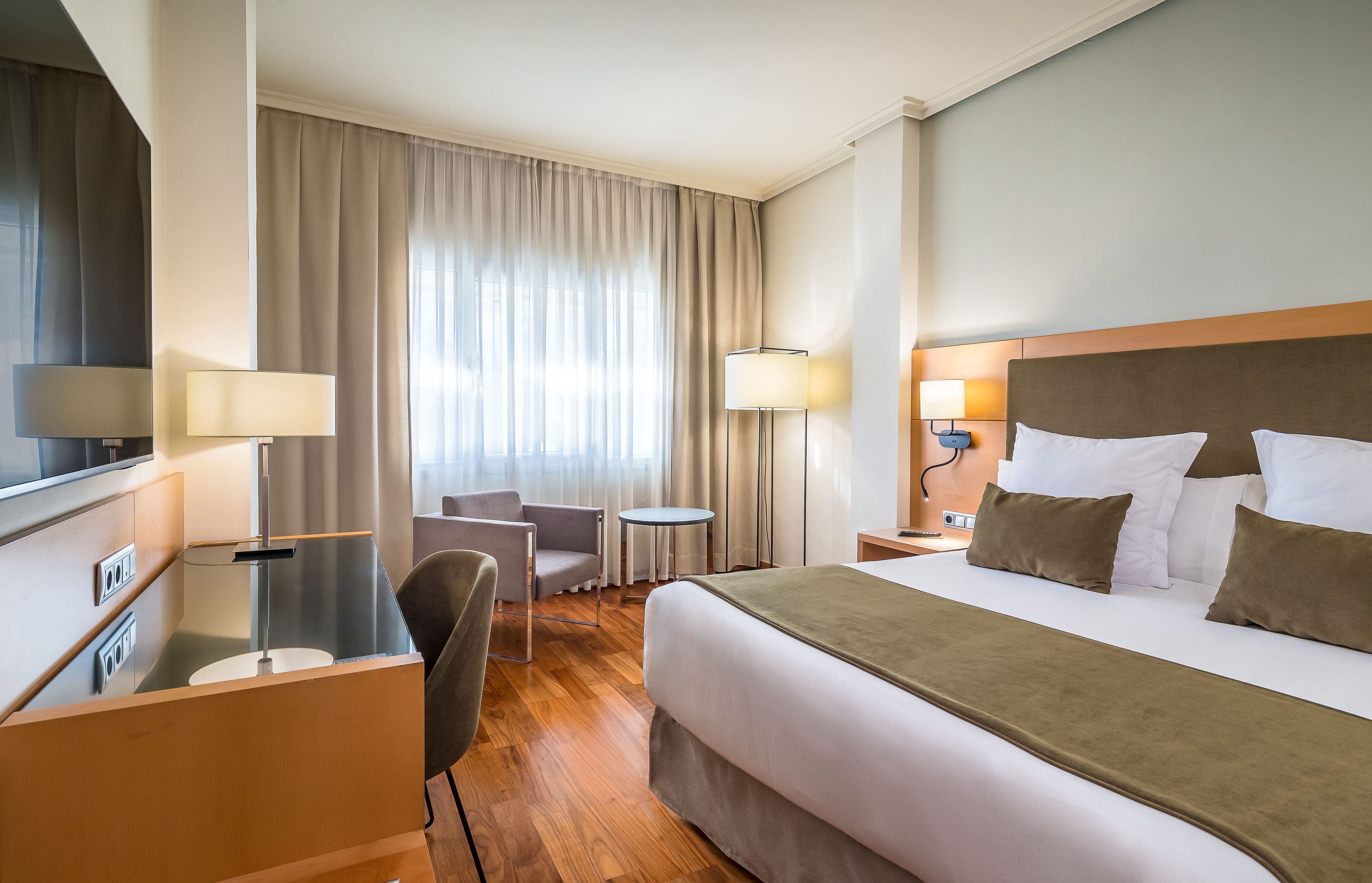 A hotel room with a bed, a desk, a chair, a small table, and a lamp.