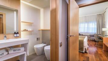 A modern hotel bathroom with a white sink, a bathtub, and a toilet. There is a wooden door leading to a bedroom with a bed and a desk.