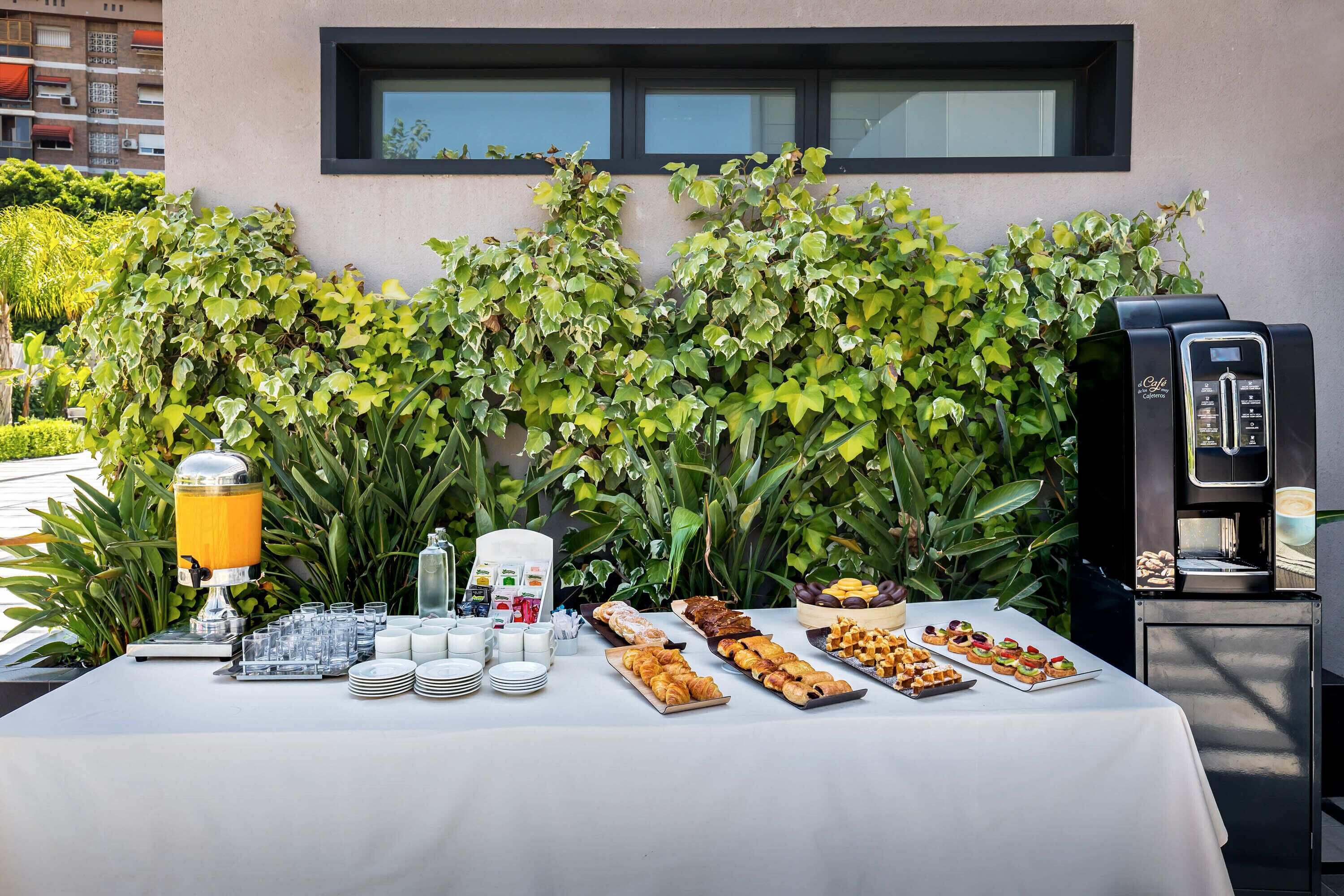 Ein reichhaltiges Buffet im Freien mit verschiedenen Speisen, einem Getränkespender und einer Kaffeemaschine vor einer Gebäudefassade.