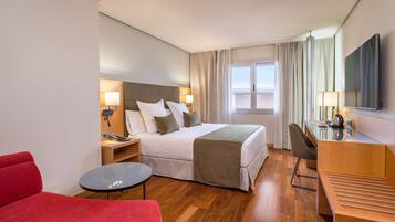 A hotel room with a bed, bedside tables, a desk, a chair, a red sofa, and a window with curtains.