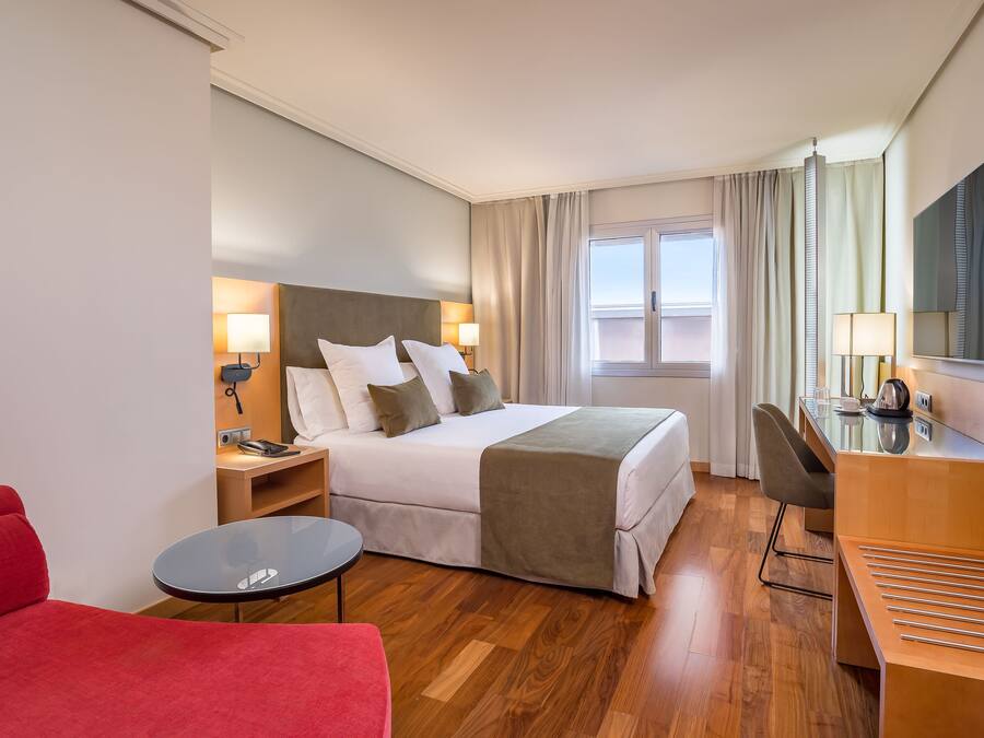 A hotel room with a bed, bedside tables, a desk, a chair, a red sofa, and a window with curtains.