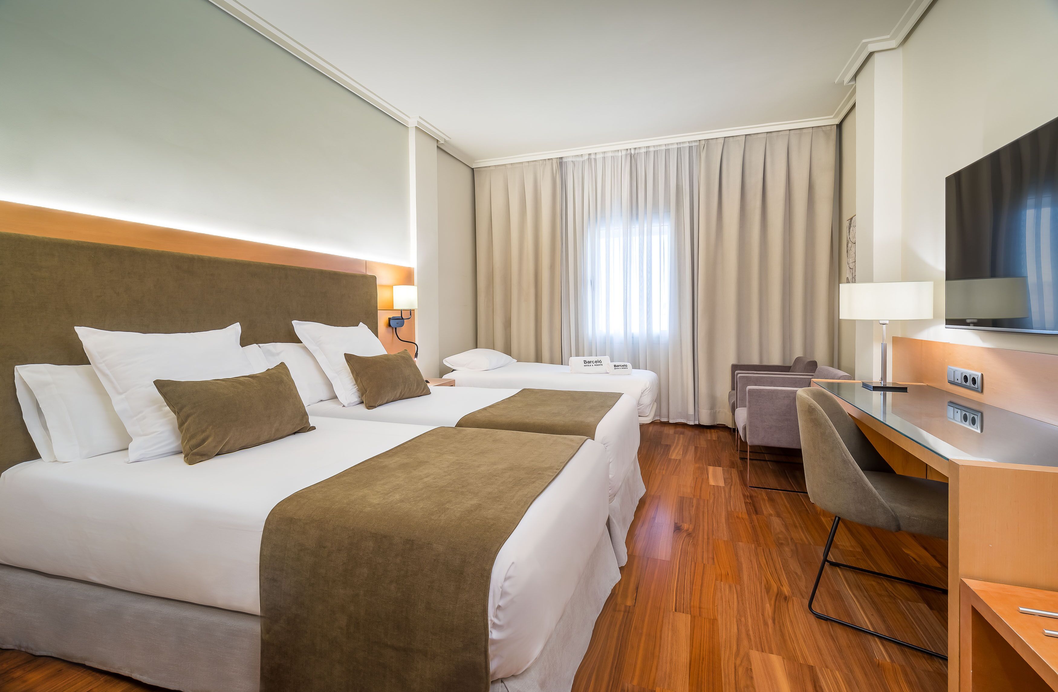 Ein Hotelzimmer mit zwei Betten, einem Schreibtisch, einem Fernseher und einem großen Fenster mit Vorhängen.