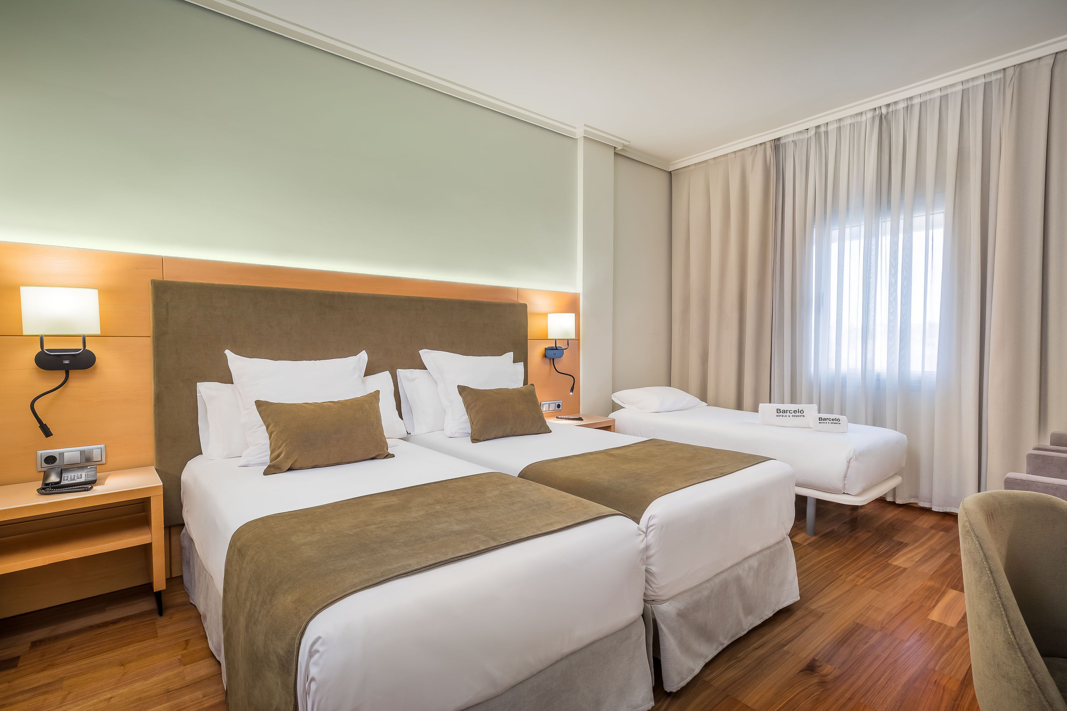 A hotel room with two beds, a wooden headboard, bedside lamps, a desk with a telephone, and a chair.