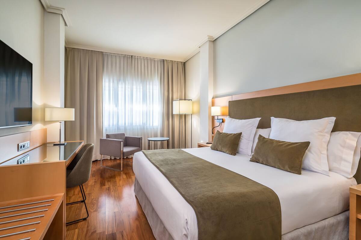Une chambre d’hôtel avec un grand lit, un bureau avec une chaise, une télévision et une fenêtre avec des rideaux.