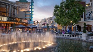Una plaza pública con una fuente, tiendas y un edificio destacado con la señal 'The Cheesecake Factory'.