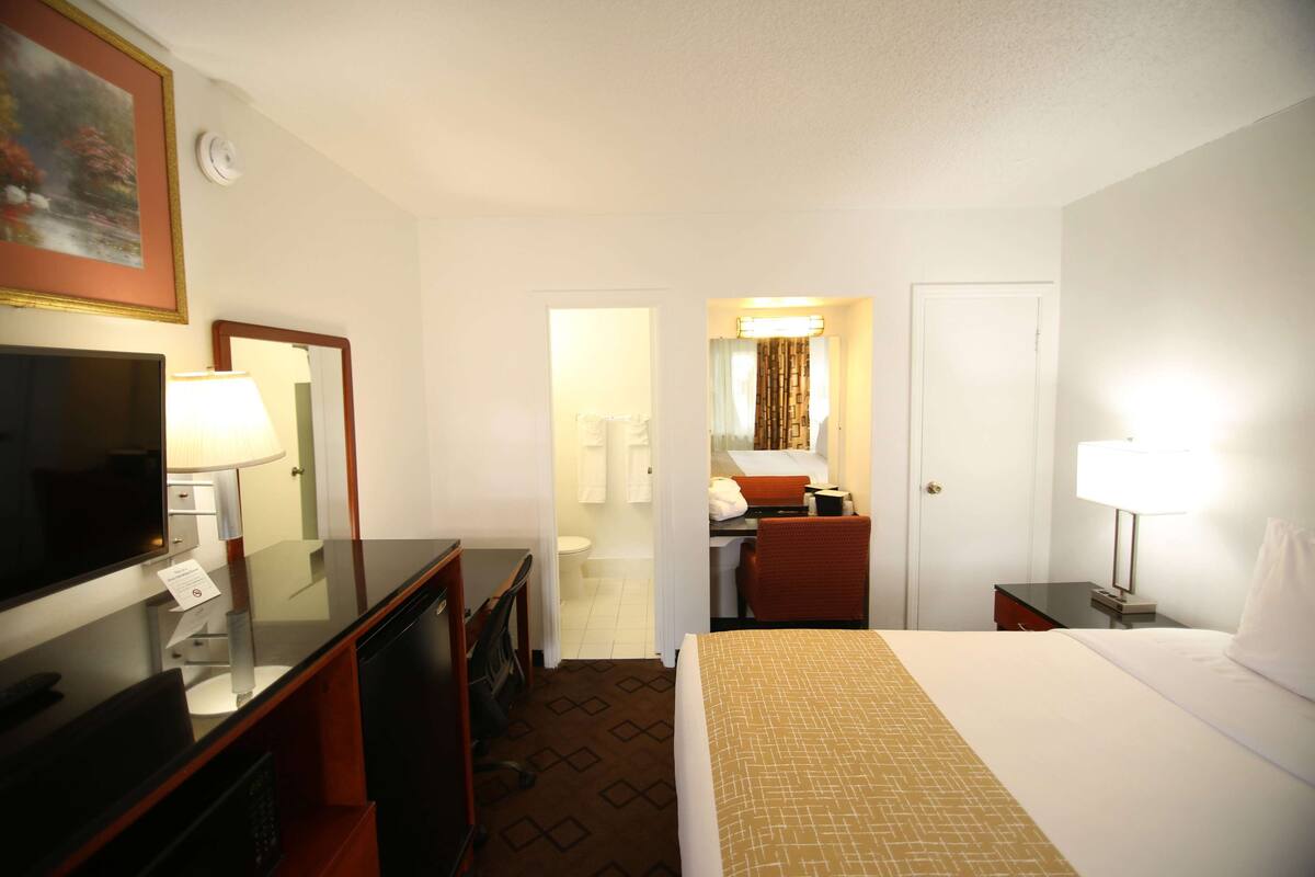 A hotel room with a bed, a desk with a computer, a TV, a mirror, and a bathroom visible through an open door.