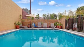 Piscina con sillones de descanso y sombrillas, rodeada por una valla de madera y plantas en macetas.