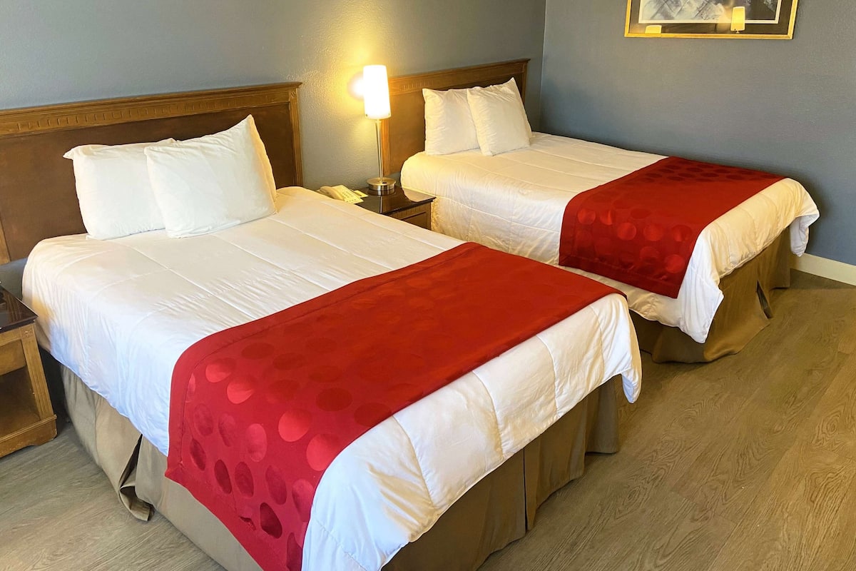 Une chambre d’hôtel avec deux lits, chacun recouvert de draps blancs et d’une couverture rouge. Une table de chevet avec une lampe et un téléphone se trouve sur le côté gauche.