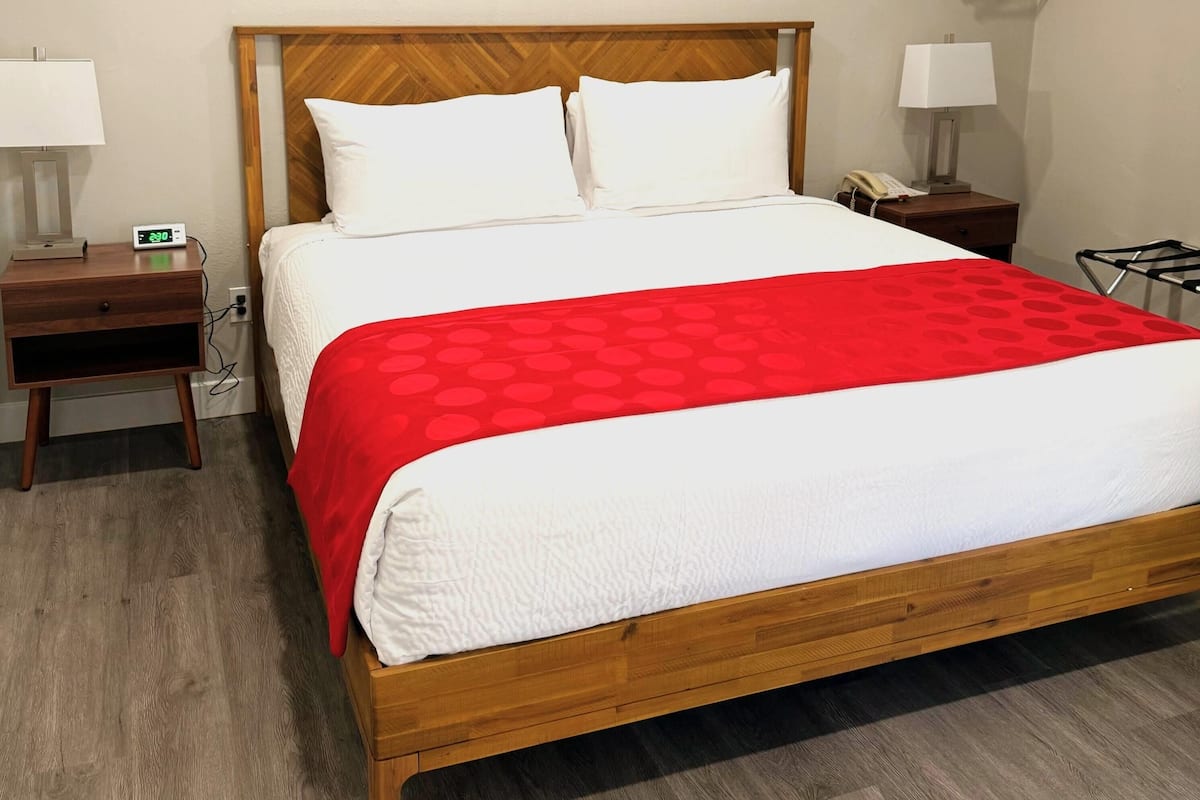 Un lit bien fait, recouvert d’une couvre-lit rouge et blanche, avec une tête de lit en bois, deux tables de chevet dotées de lampes et une petite table de nuit avec un téléphone.