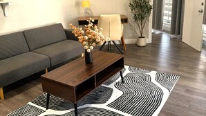 Een moderne woonkamer met een donkerbruine salontafel, een grijze bank, een zwart-wit tapijt met patroon, een potplant en een lamp op een bijzettafeltje.
