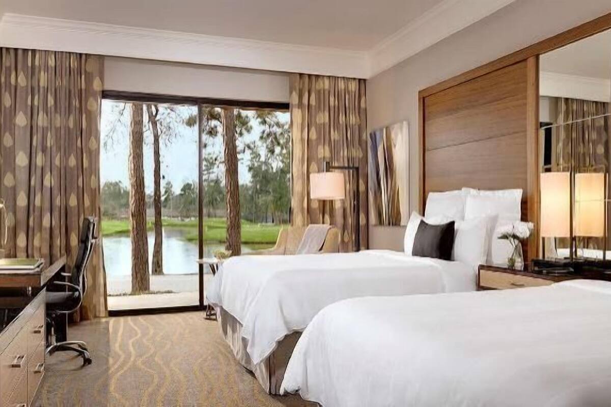 A hotel room with two beds, a desk, a chair, a TV, and a large window with a view of trees and a body of water.