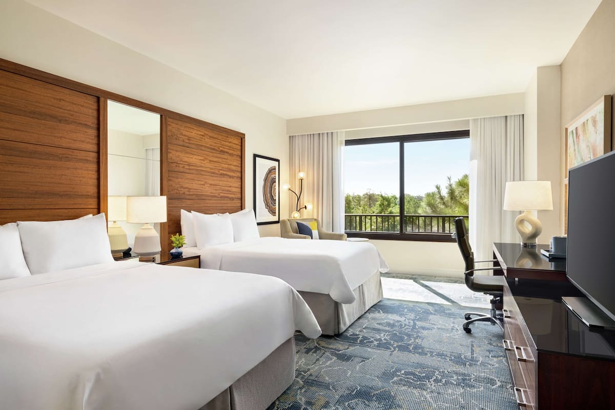 A hotel room with two beds, a desk with a TV, a large window with a view of trees, and a balcony.