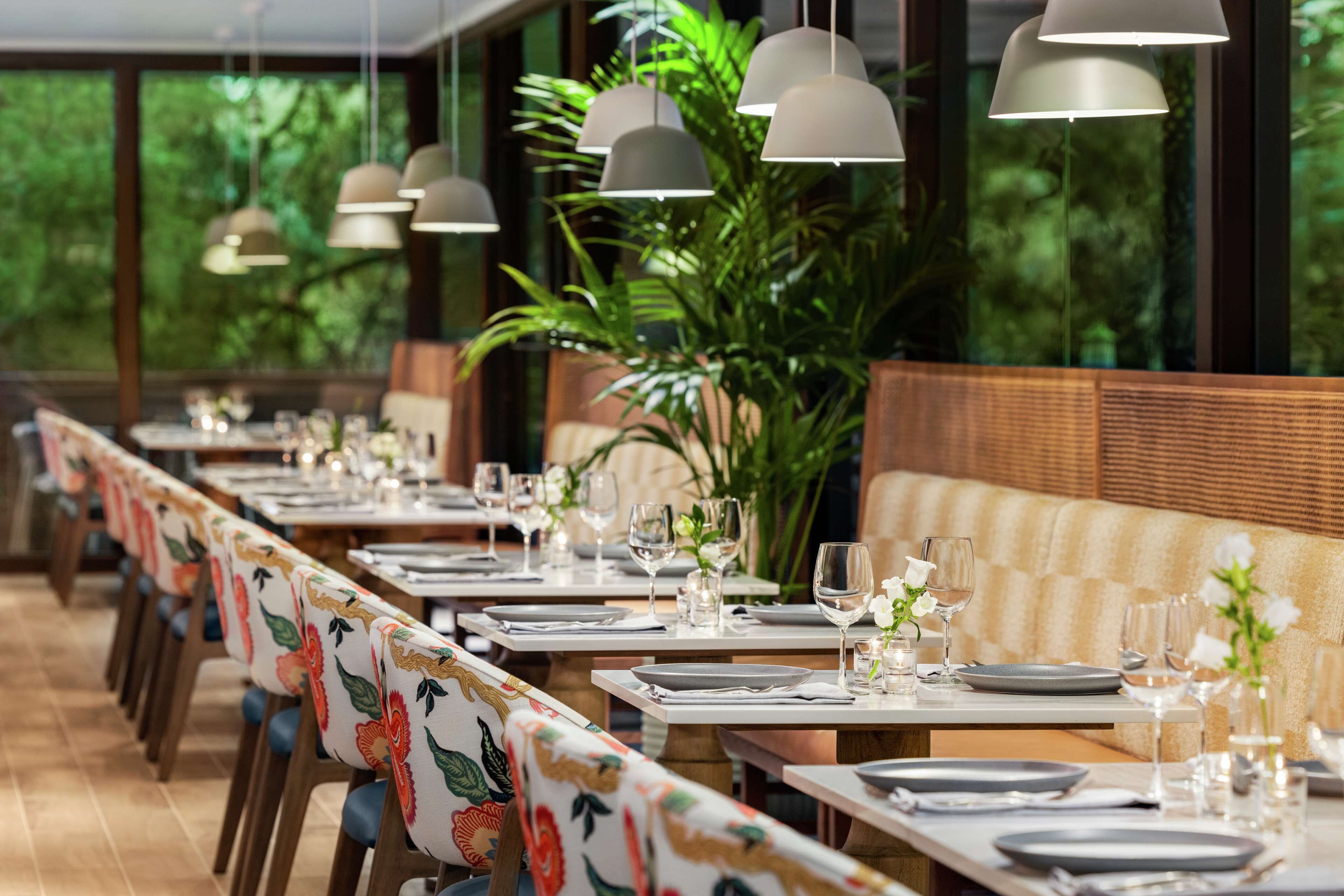 Un restaurante con mesas largas, sillas con tapicería floral, lámparas colgantes y vistas a zonas verdes a través de amplios ventanales.