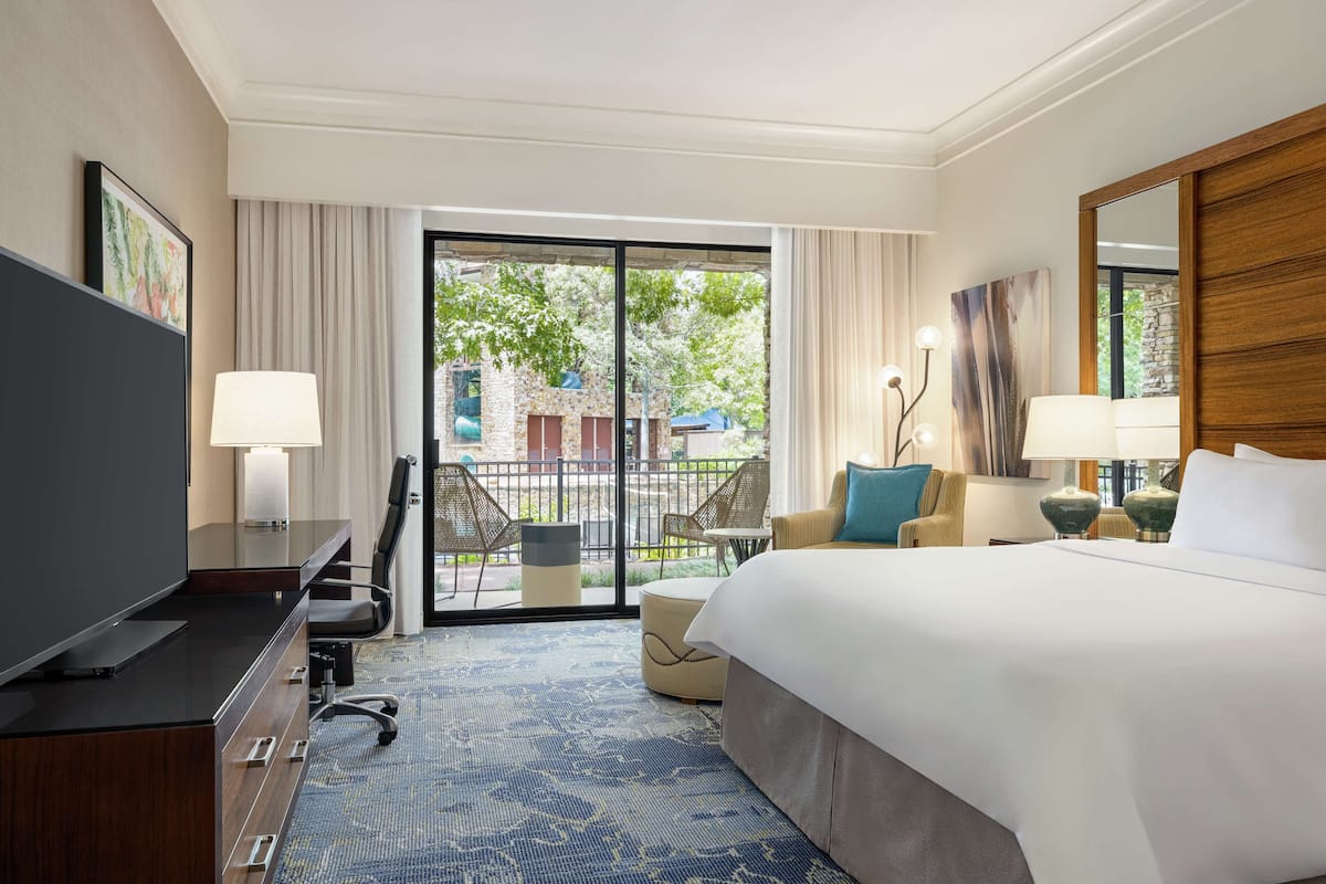 A hotel room with a large bed, a desk with a TV, a chair, a small table, and a balcony with chairs and a view of the outside.