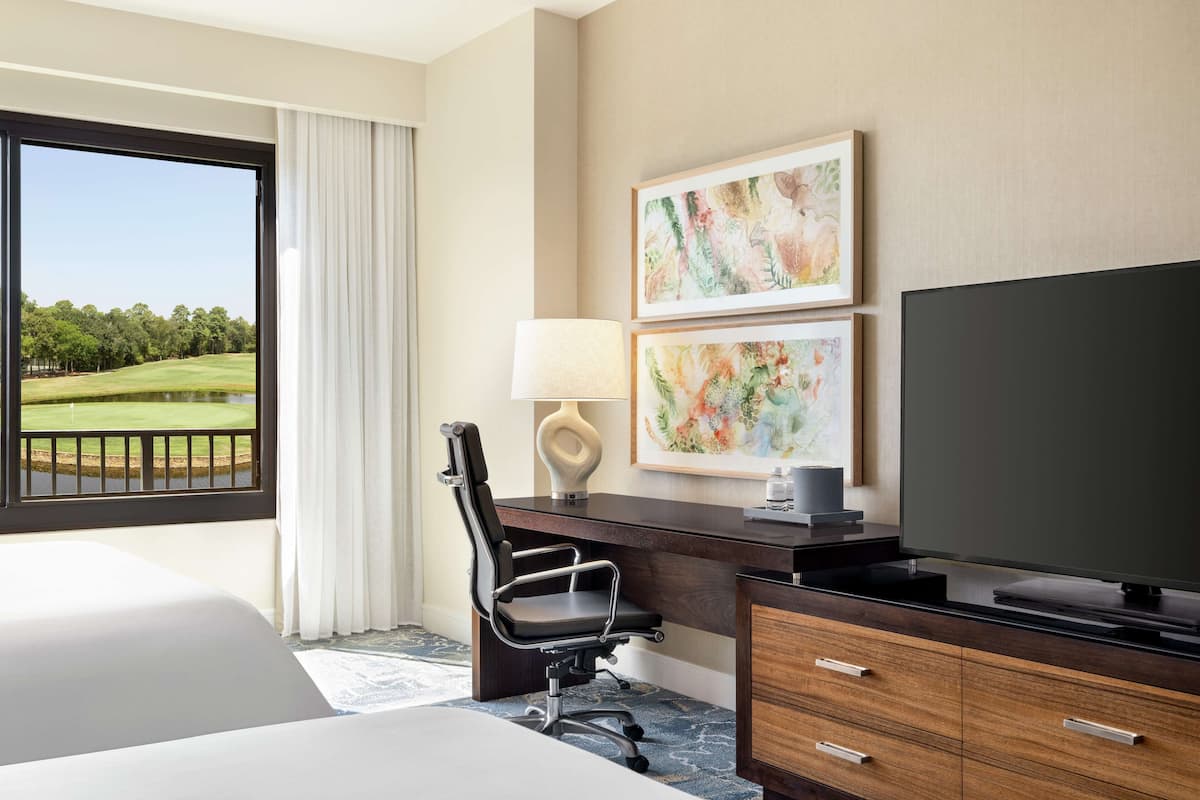 A hotel room with a large bed, a desk with a chair, a television, and two framed artworks.