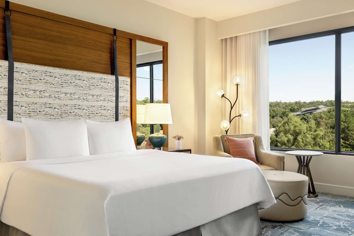 A hotel room with a large bed, a wooden headboard, a view of greenery outside, a small round table, and a chair.