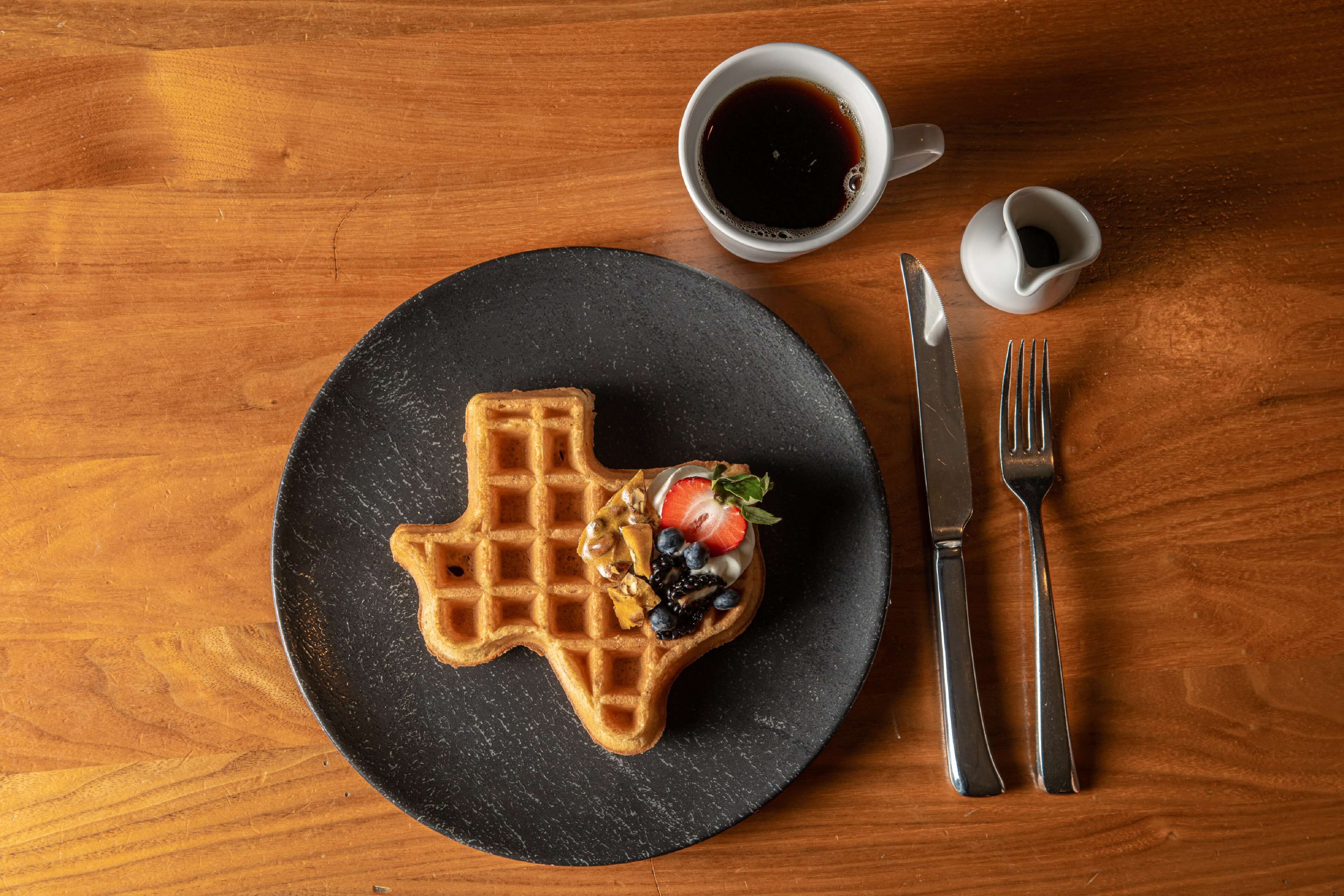 A waffle on a black plate with a cup of coffee and a small cup of syrup.