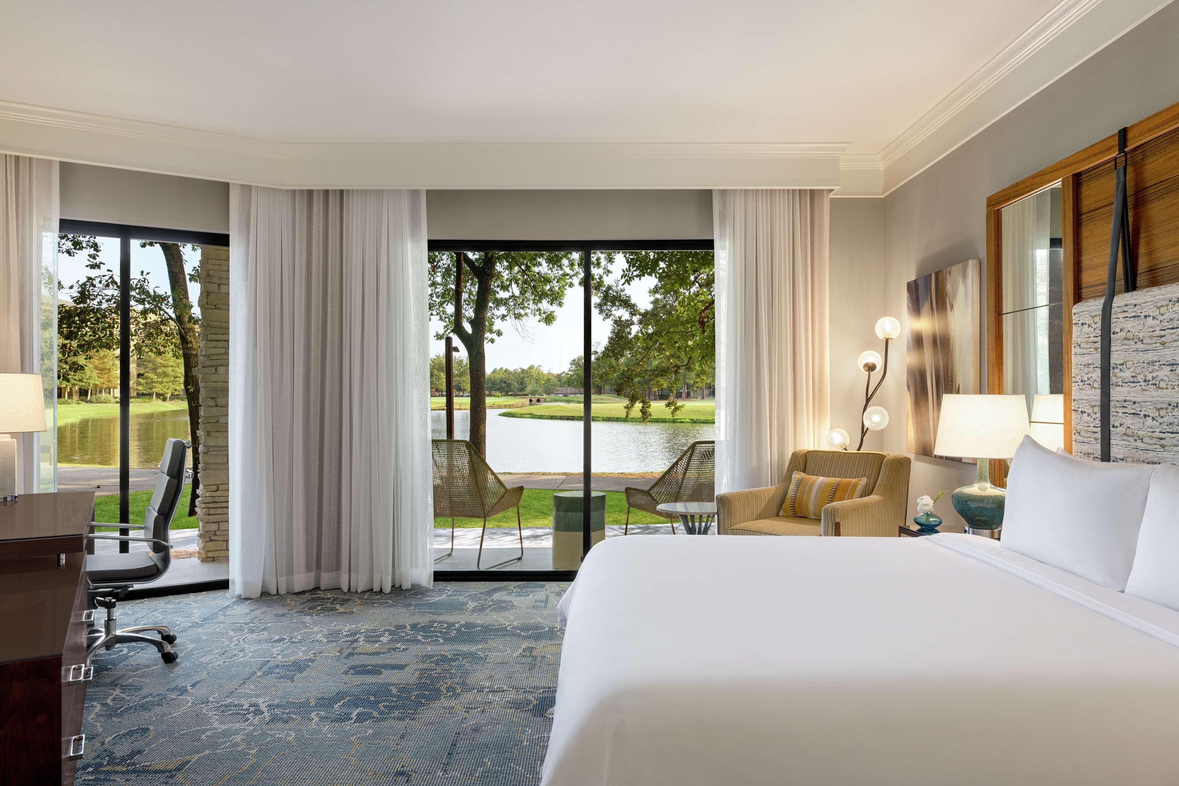 Une chambre d’hôtel dotée d’un grand lit, d’un bureau et offrant une vue sur un lac et un parc grâce à de grandes fenêtres du sol au plafond.