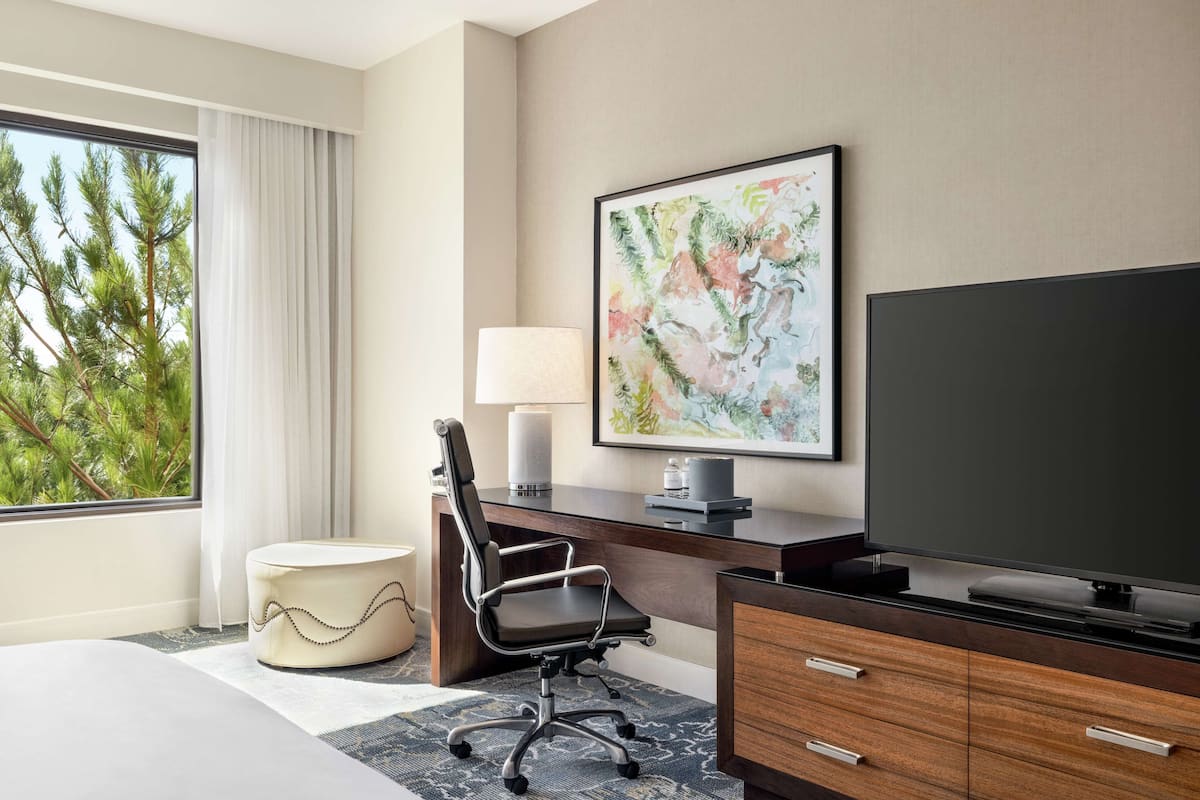 A hotel room with a large window, a desk with a chair, a television, and a bed.