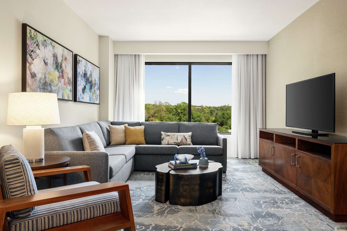 A modern hotel room with a grey sofa, a wooden coffee table, a TV on a wooden cabinet, and a large window with a view of greenery.