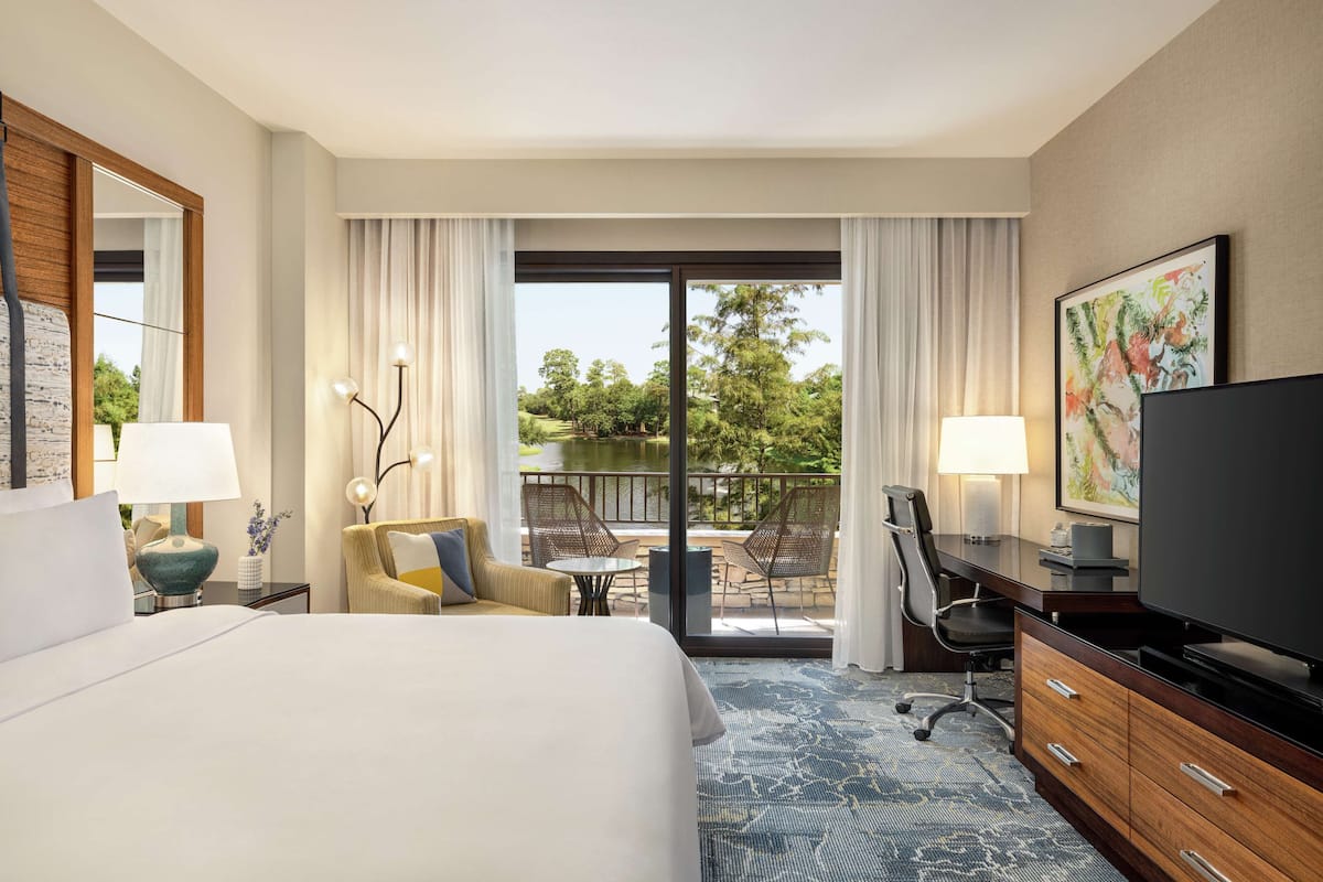 A hotel room with a large bed, a desk with a television, a chair, a small table, and a balcony with a view of trees and a body of water.