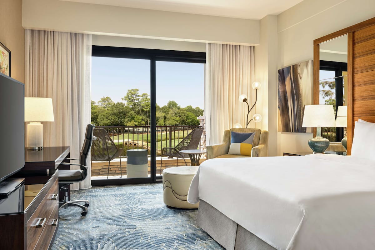 A hotel room with a large bed, a desk with a TV, a chair, a small table, and a balcony with a view of trees.