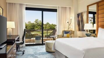 Habitación de hotel con una cama grande, un escritorio con televisor, una silla, una mesita y un balcón con vistas a árboles.
