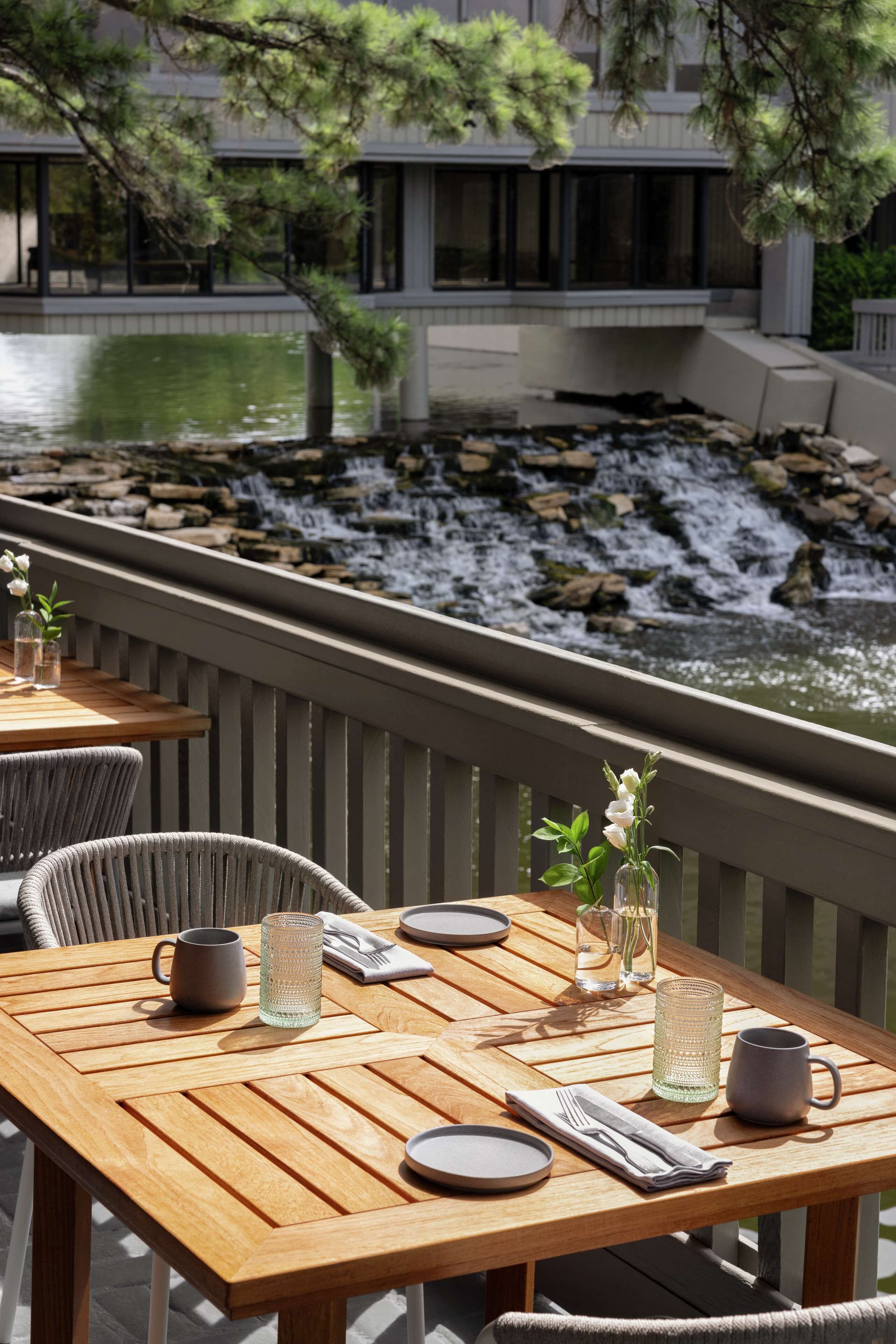 Mesa de madera dispuesta con platos, vasos y cubiertos, con vista a una cascada y follaje.
