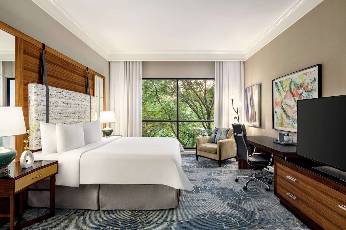 A hotel room with a large bed, a desk, a chair, a television, and a window with a view of trees.
