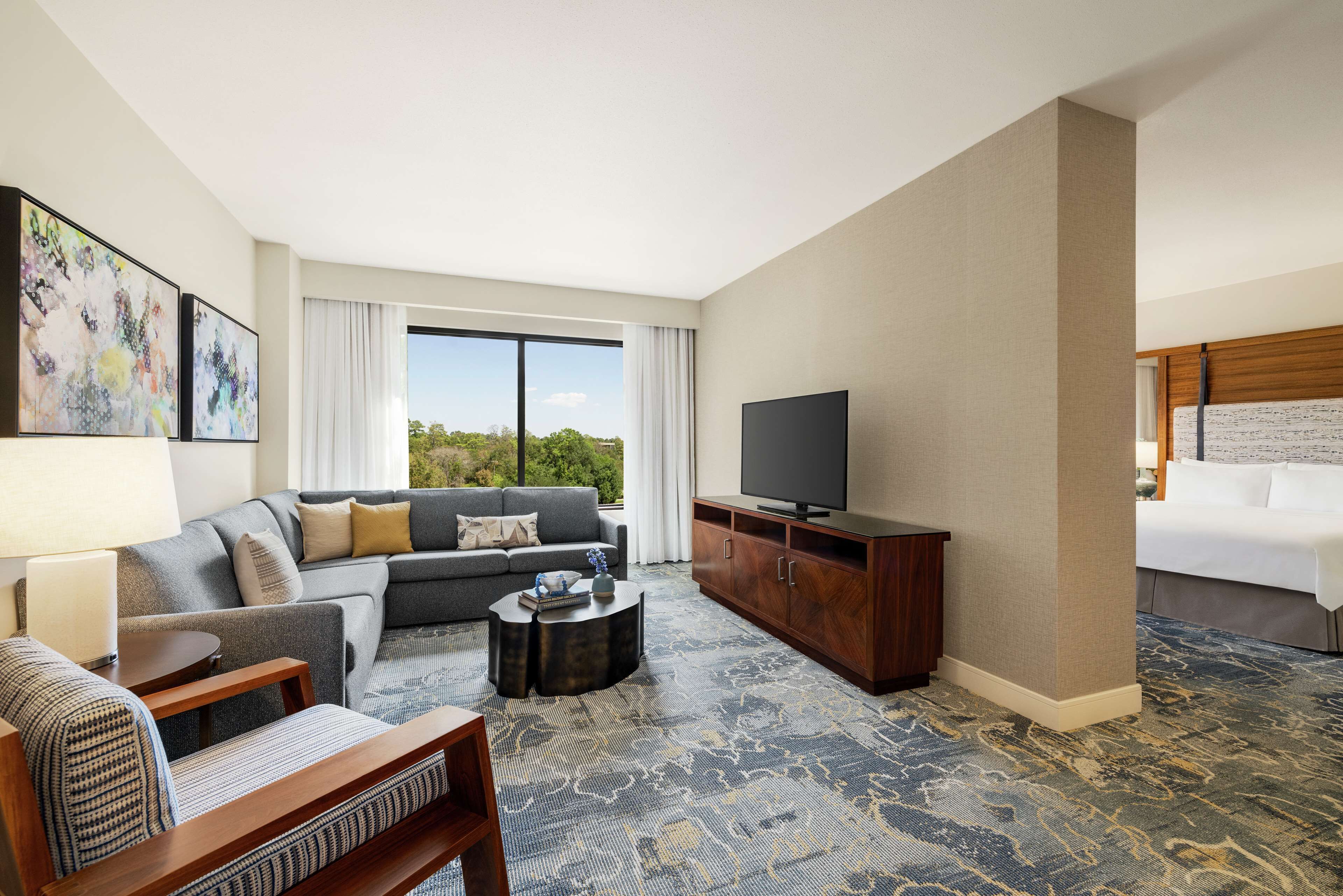 A hotel room with a sofa, a TV, a wooden cabinet, a patterned carpet, and a large window with a view of greenery.