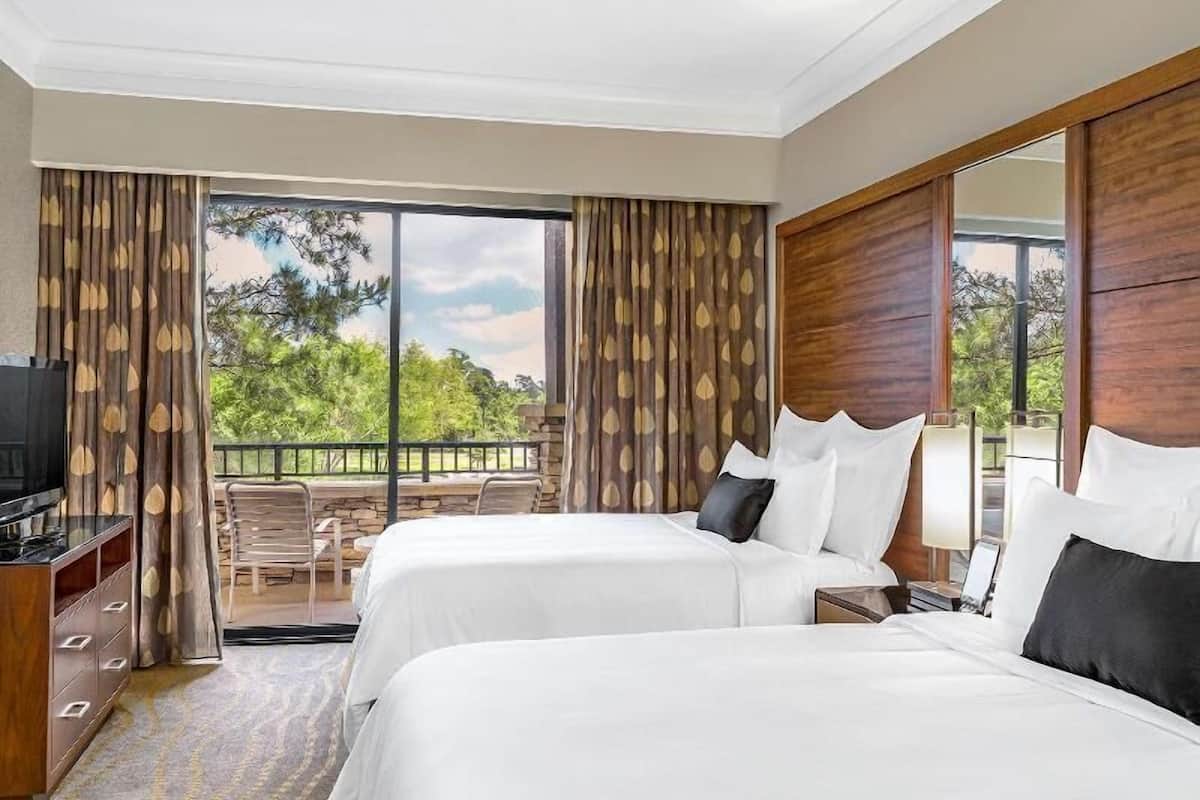 A hotel room with two beds, a TV, a desk, and a balcony with a view of trees.