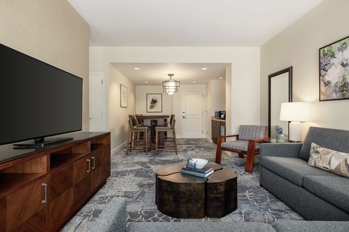 A modern living room with a flat-screen TV, a grey sofa, a round coffee table, and a dining area with a chandelier.