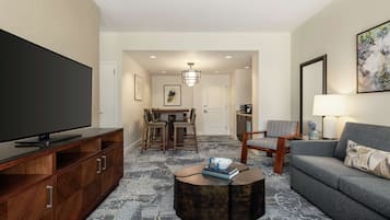 A modern living room with a flat-screen TV, a grey sofa, a round coffee table, and a dining area with a chandelier.
