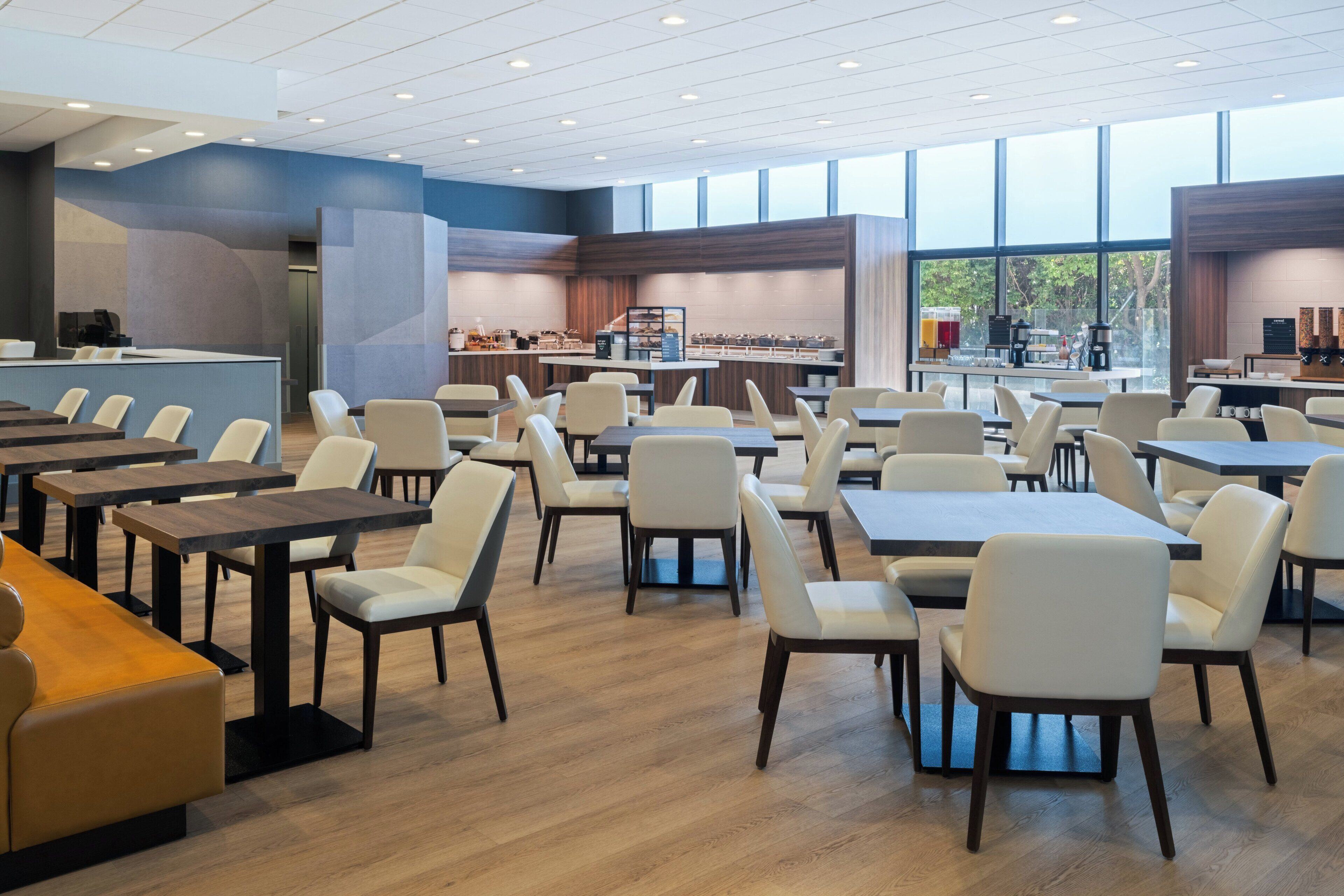 Una amplia cafetería con numerosas mesas y sillas, una estación de café y vistas al exterior gracias a grandes ventanales.