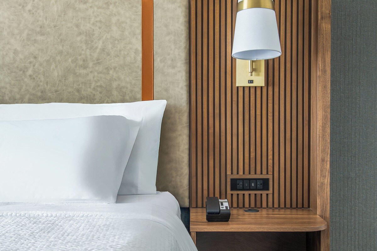 Quarto de hotel com uma cama, uma mesa de cabeceira com abajur e parede de painéis de madeira.