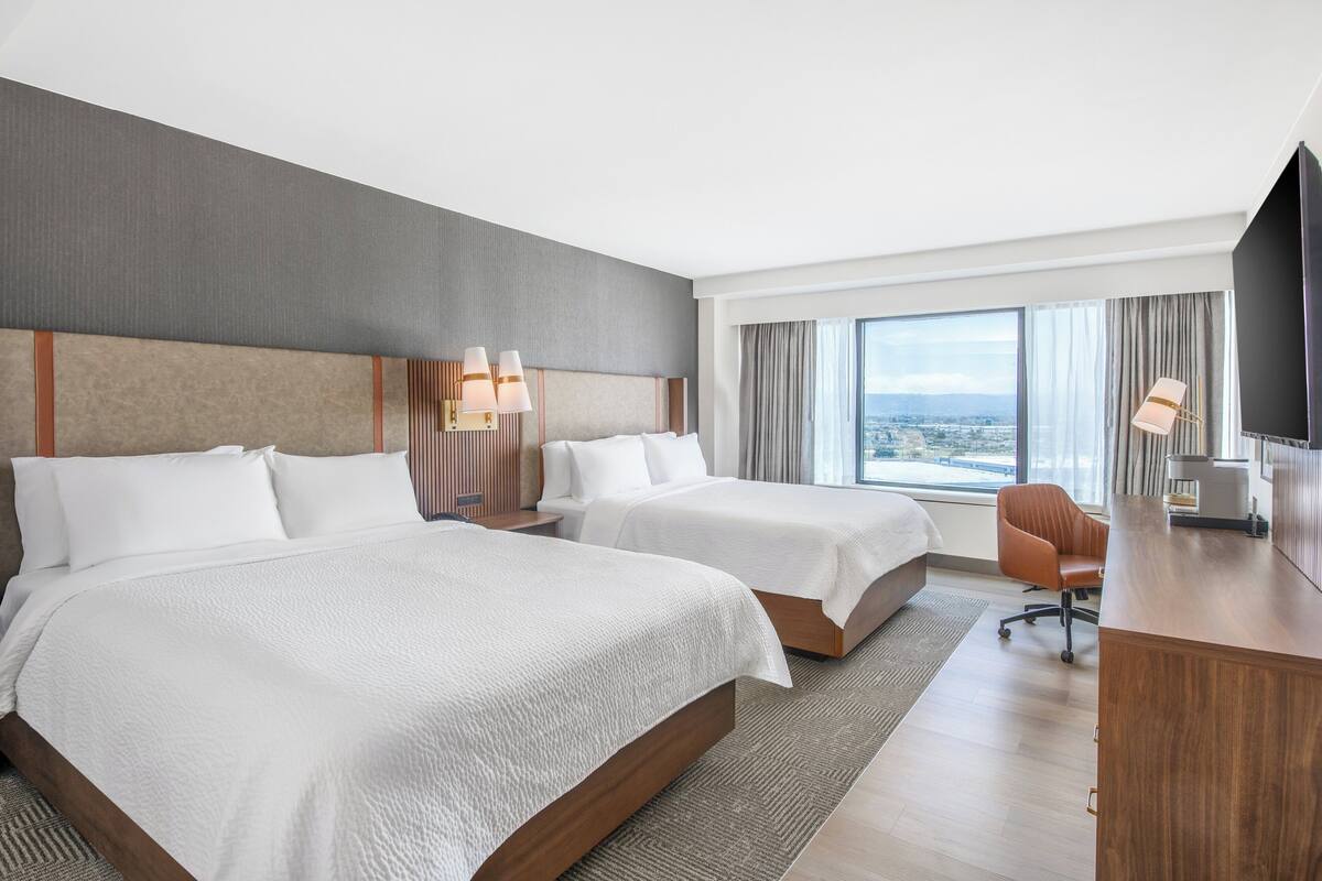 A hotel room with two beds, a desk, and a large window offering a city view.