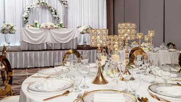 A formal dining setting with tables set for a meal, featuring gold and white tableware, clear glassware, and elegant lighting fixtures.