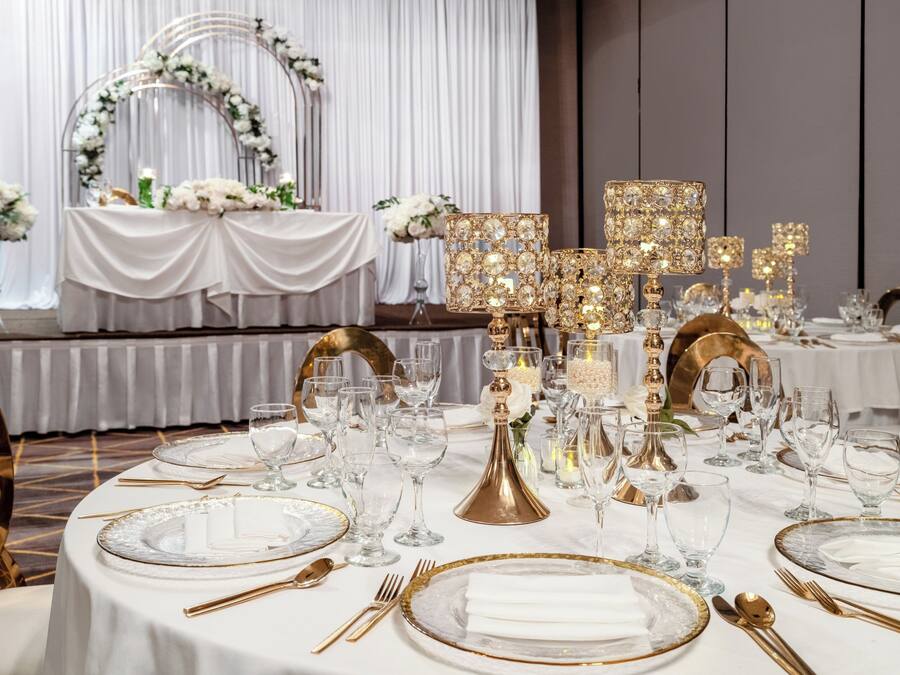 A formal dining setting with tables set for a meal, featuring gold and white tableware, clear glassware, and elegant lighting fixtures.