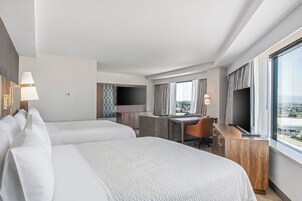 A hotel room with two beds, a desk, a TV, and a view of the city.