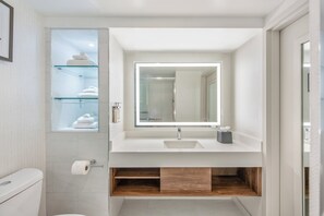 Une salle de bain moderne avec un meuble-lavabo blanc, une étagère en verre où sont posées des serviettes et un miroir doté d’un éclairage intégré.
