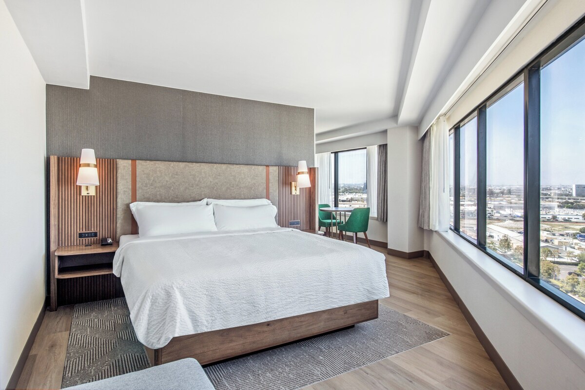 Quarto de hotel moderno com uma cama grande, duas mesas de cabeceira com luminárias, uma escrivaninha pequena, uma cadeira e uma janela ampla com vista para a cidade.