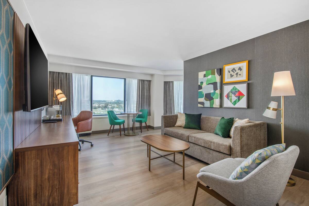 A modern hotel room with a wooden desk, a sofa, two armchairs, a coffee table, and a TV. There are large windows with curtains, a city view, and artwork on the walls.