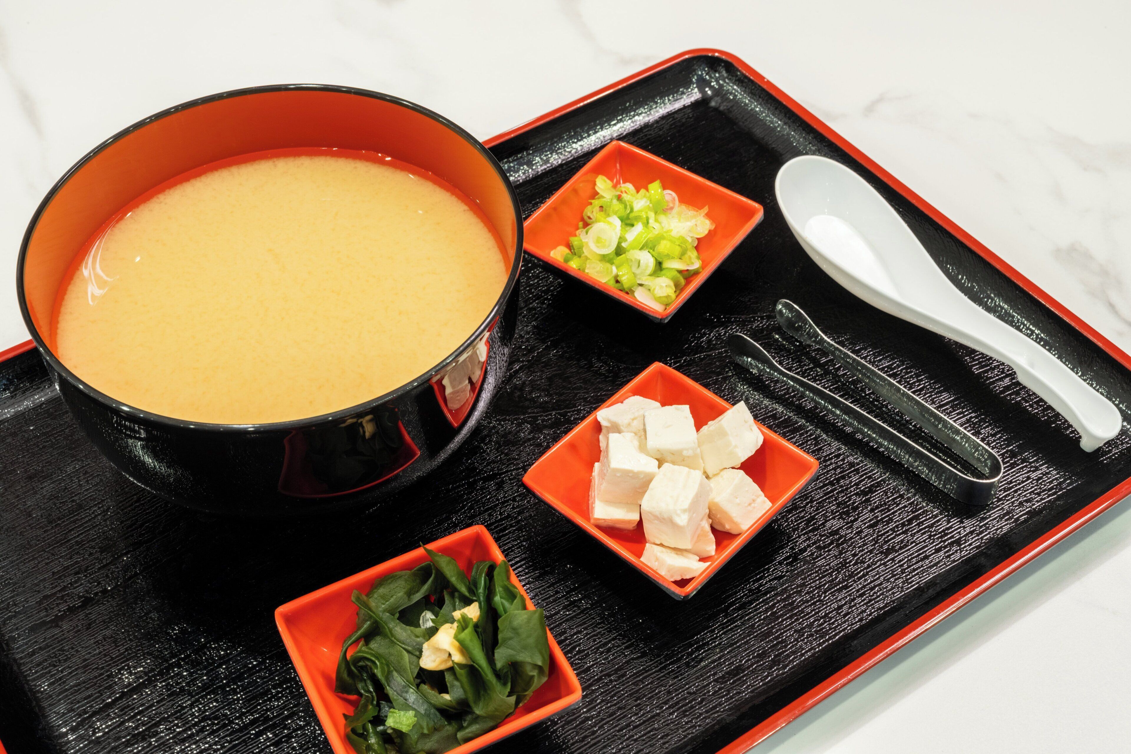 A tray with a bowl of soup, a small bowl of chopped vegetables, a small bowl of tofu, and a pair of chopsticks.