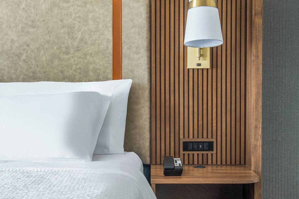 A hotel room with a bed, a nightstand, a lamp, and a wall-mounted control panel.