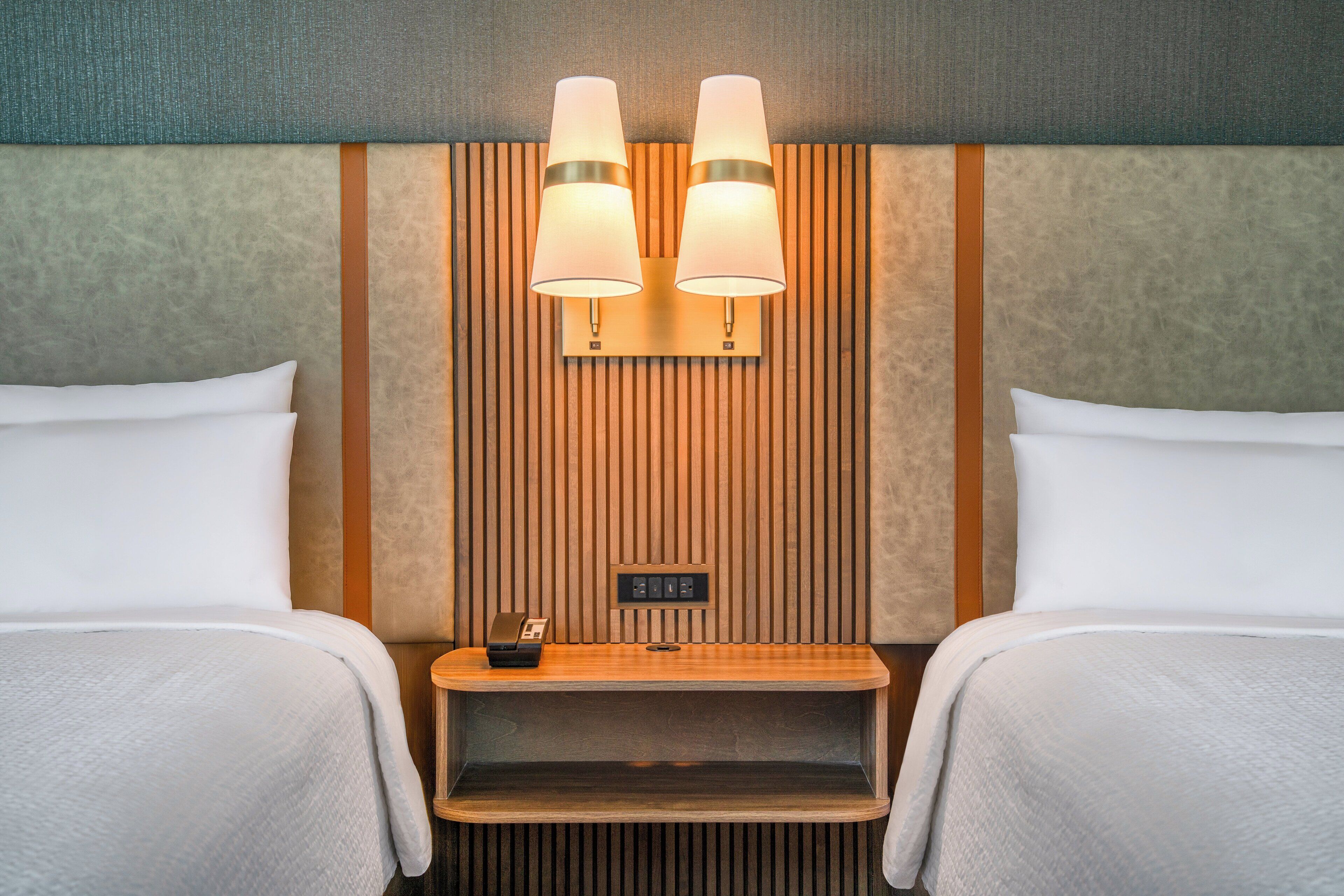 Une chambre d’hôtel avec deux lits, une table de chevet en bois avec un téléphone et une petite lampe, et un luminaire fixé au mur.