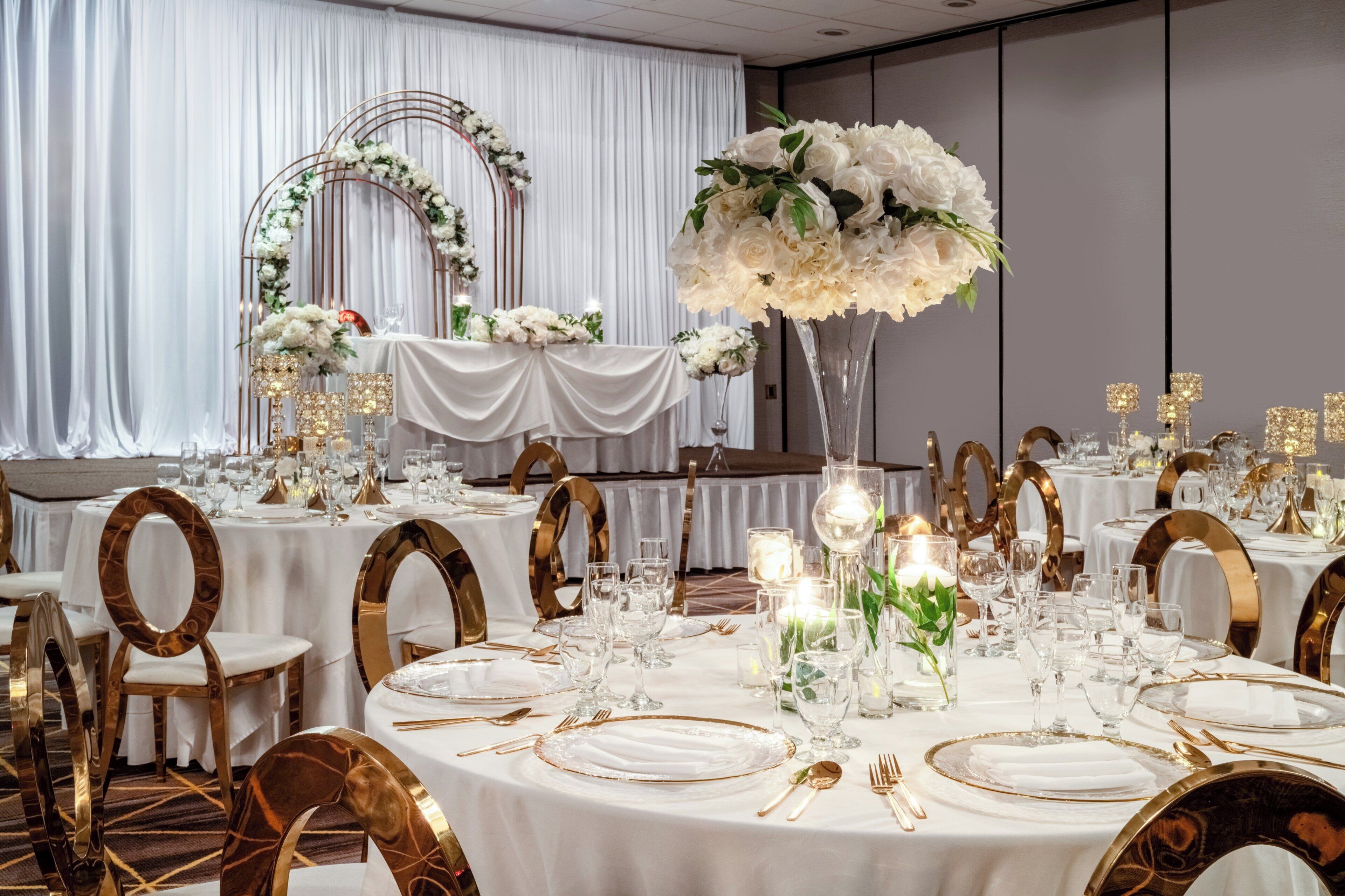 Une salle de banquet avec des tables rondes dressées pour un événement formel, agrémentée de nappes blanches, de touches dorées et de compositions florales en guise de centre de table.