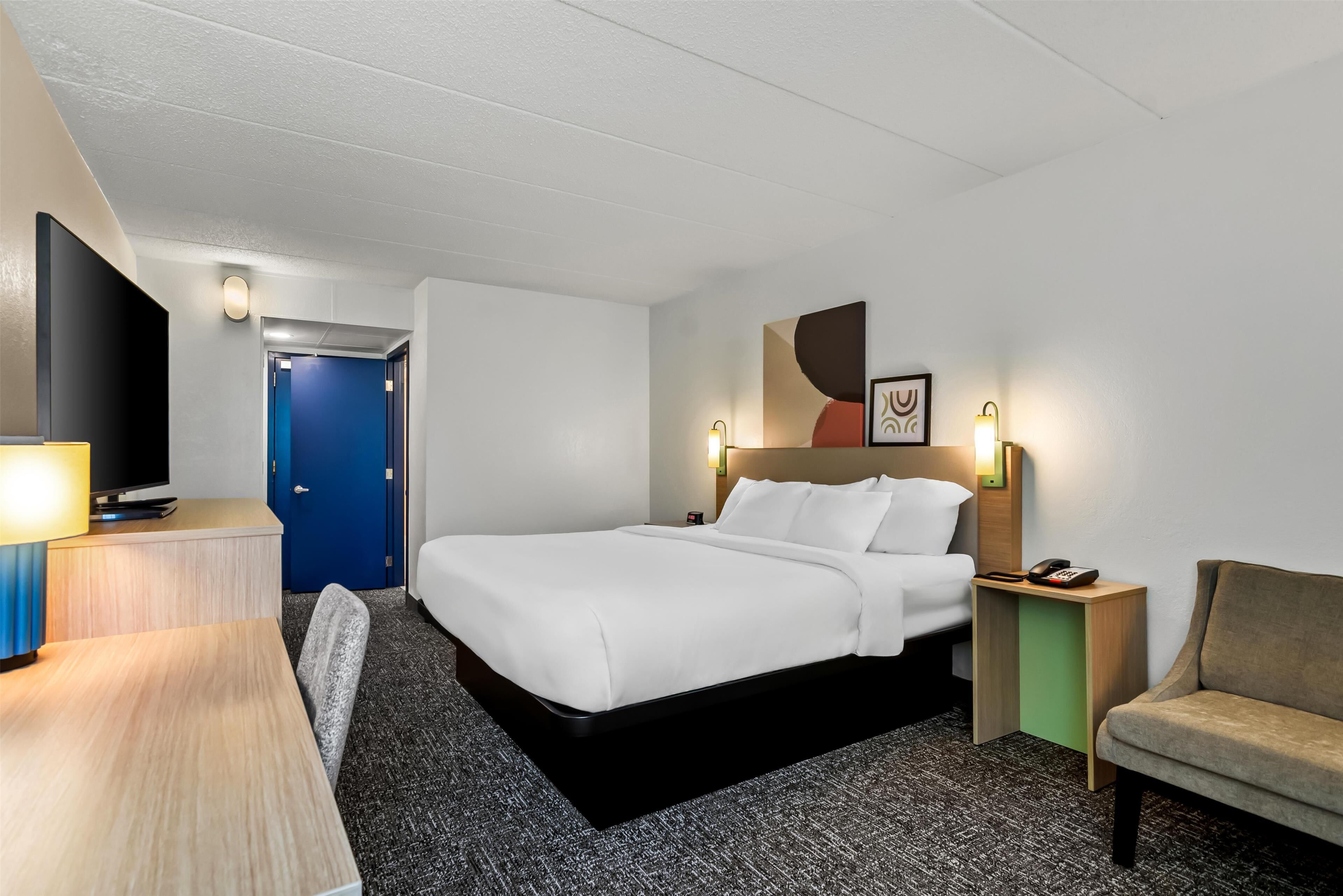 A hotel room with a bed, bedside table, lamp, TV, and a chair.